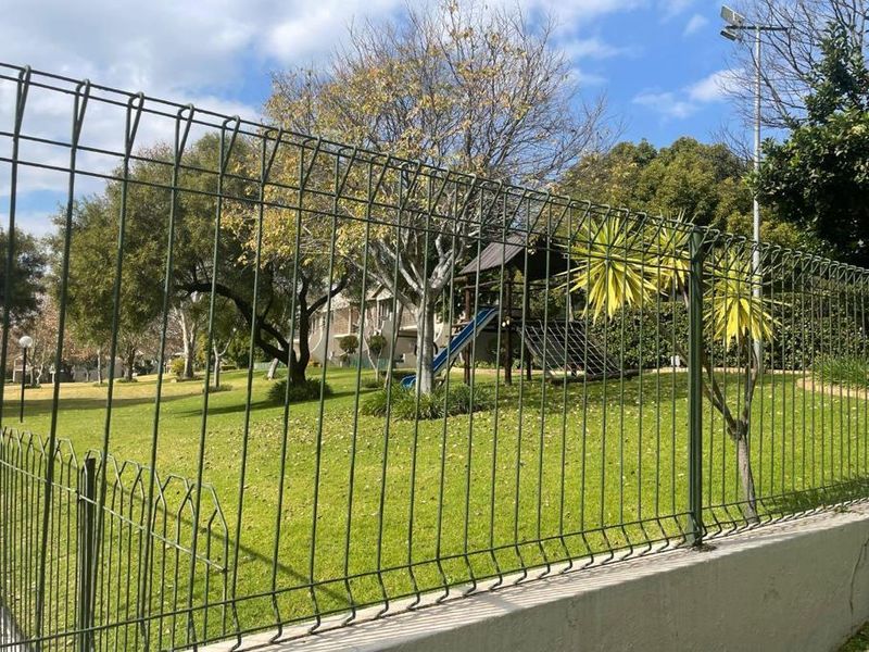 A well-maintained communal garden area featuring a lawn, mature trees, and a children's playground, secured by a green mesh perimeter fence and retaining wall.