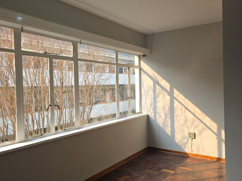 A bright, empty living area featuring classic parquet flooring, large windows allowing natural light, and modern track lighting.