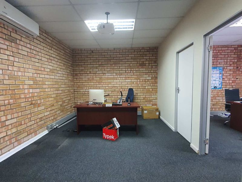 A functional office space featuring exposed brick walls, carpeted flooring, a suspended ceiling, and an integrated air conditioning unit.