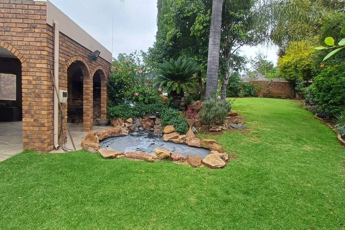 A well-maintained rear garden featuring a brick-arched patio, a landscaped rock-lined water feature, and a spacious lawn area.