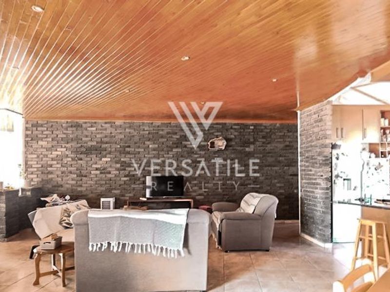 Open-plan living area featuring a distinctive timber-clad vaulted ceiling, exposed brick feature wall, and tiled flooring, seamlessly connecting to the kitchen space.
