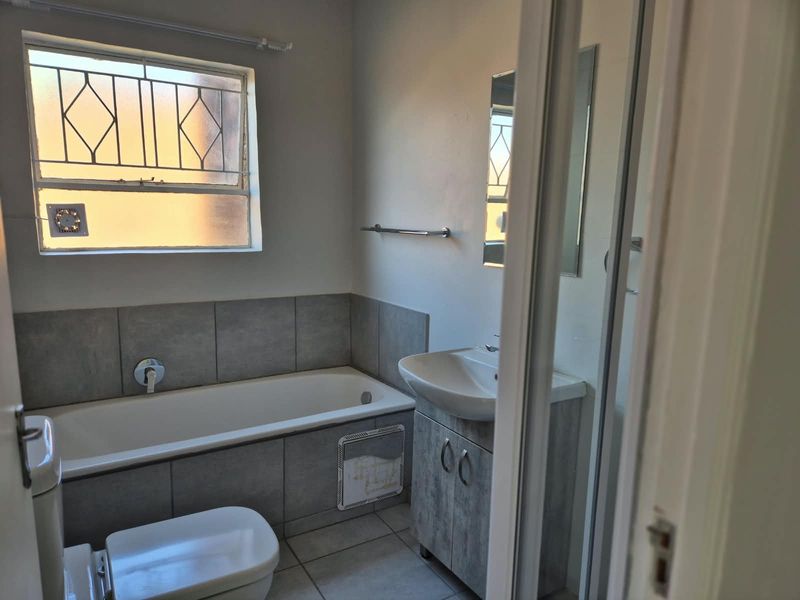 A functional bathroom featuring a bathtub, vanity cabinet with basin, and toilet, finished with neutral wall tiles and a window with security bars.
