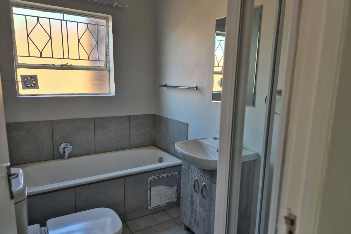 A functional bathroom featuring a bathtub, vanity cabinet with basin, and toilet, finished with neutral wall tiles and a window with security bars.
