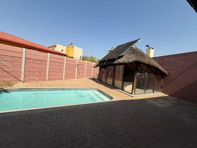 Outdoor pool area featuring a swimming pool, paved patio, and a thatched lapa with solar heating panels, enclosed by a high boundary wall.