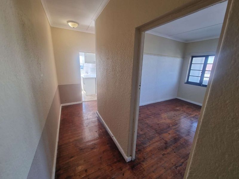 A vacant hallway with original hardwood flooring leading into an adjacent bedroom and kitchen area, featuring neutral wall tones and coved ceilings.