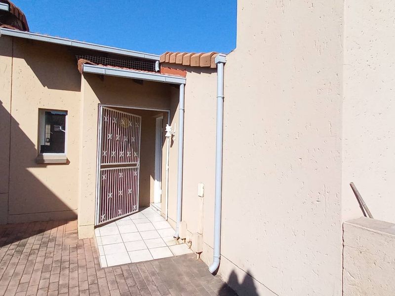 Front entrance of the townhouse featuring a security gate, paved walkway, and tiled entry area with exterior plaster finish.