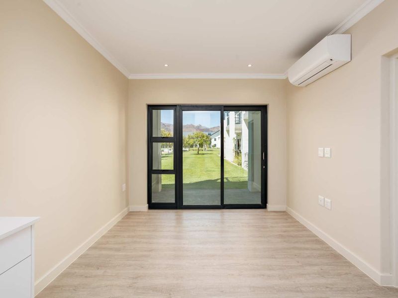 A bright, empty living area featuring modern laminate flooring, recessed lighting, a wall-mounted air conditioning unit, and sliding glass doors leading to the garden.