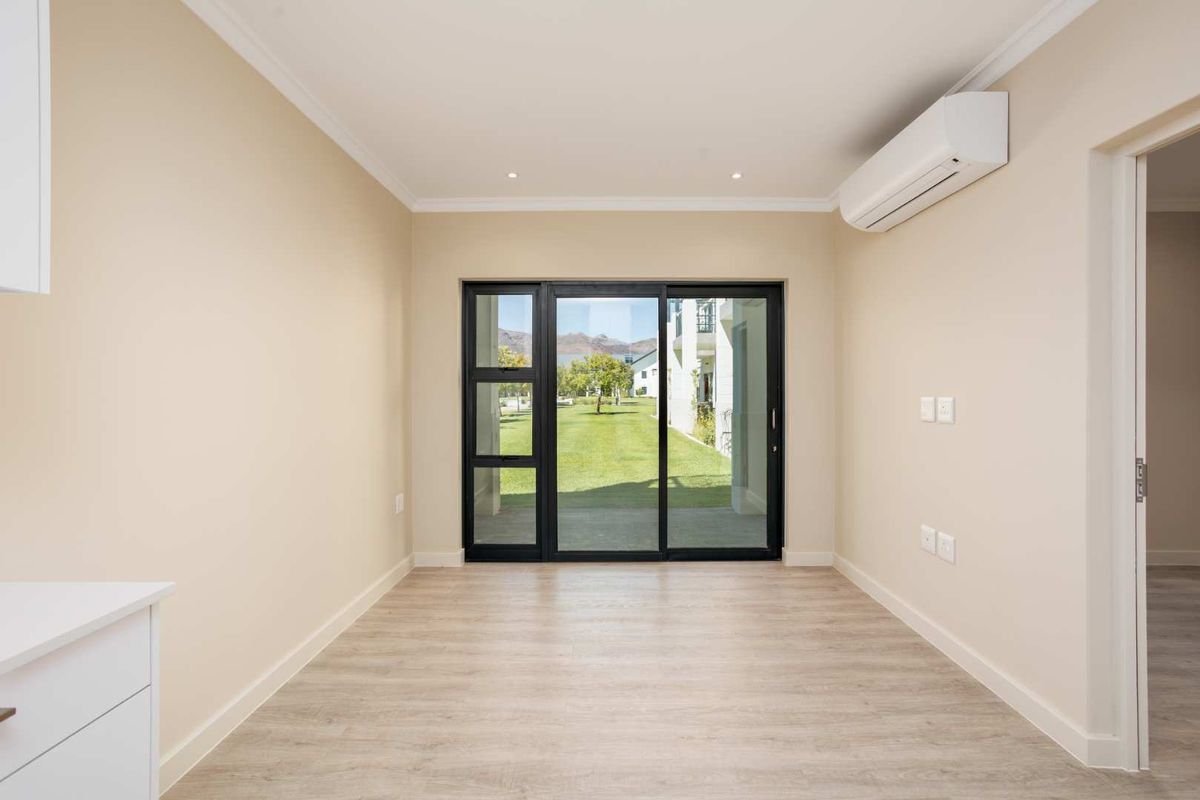 A bright, empty living area featuring modern laminate flooring, recessed lighting, a wall-mounted air conditioning unit, and sliding glass doors leading to the garden.