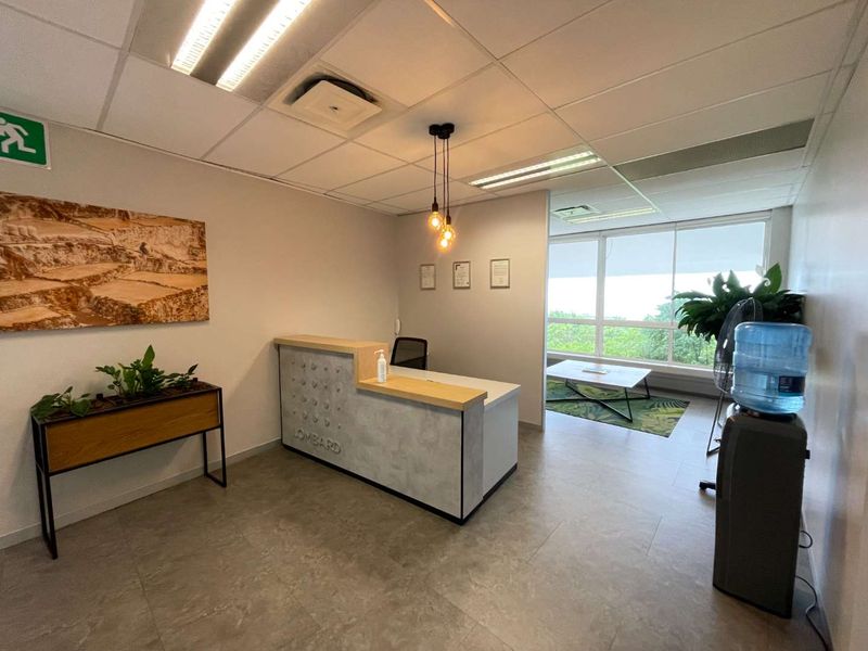 A professional office reception area featuring a modern built-in desk, tiled flooring, suspended ceiling, and large windows providing natural light.