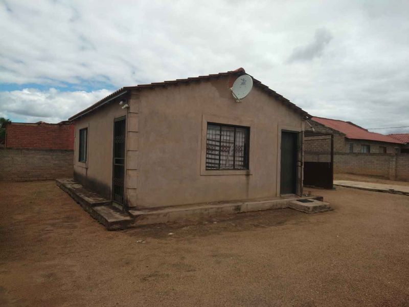 A detached single-story house featuring a tiled roof, security gates, and burglar bars, situated on a large, unpaved plot.