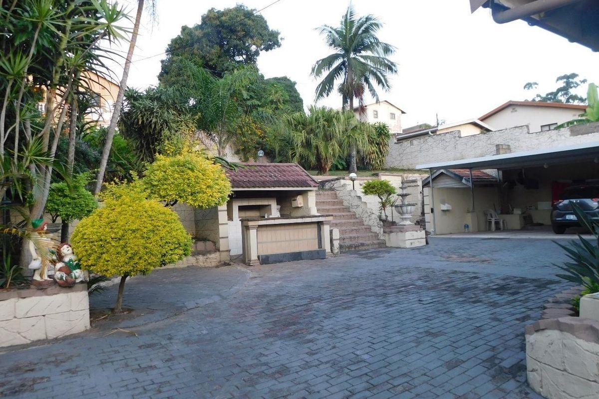 Rear exterior area featuring a paved driveway, carport, built-in braai, and stone stairs leading to a landscaped garden level.