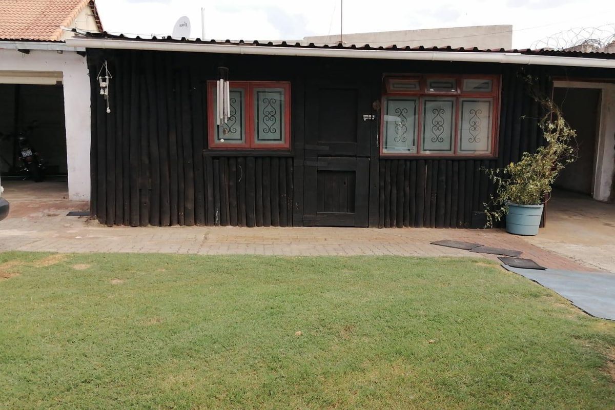 A rustic exterior view featuring dark timber log-style cladding, framed windows with security bars, and a paved area adjacent to a lawn.
