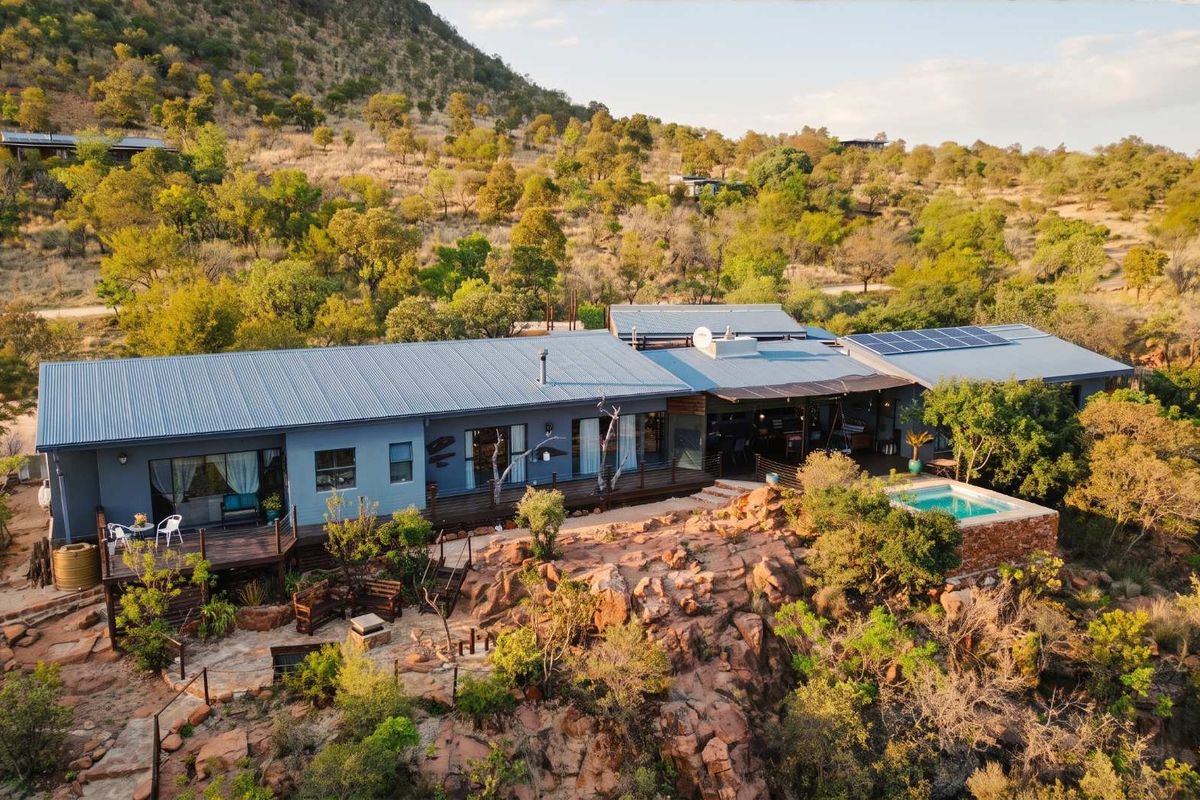 Aerial view of a modern bushveld residence featuring a corrugated metal roof, solar panels, timber decking, and a private swimming pool set in rocky terrain.