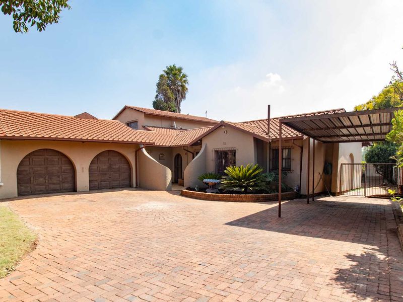 Front elevation of a house featuring a double garage, a carport, and a spacious brick-paved driveway with established garden beds.