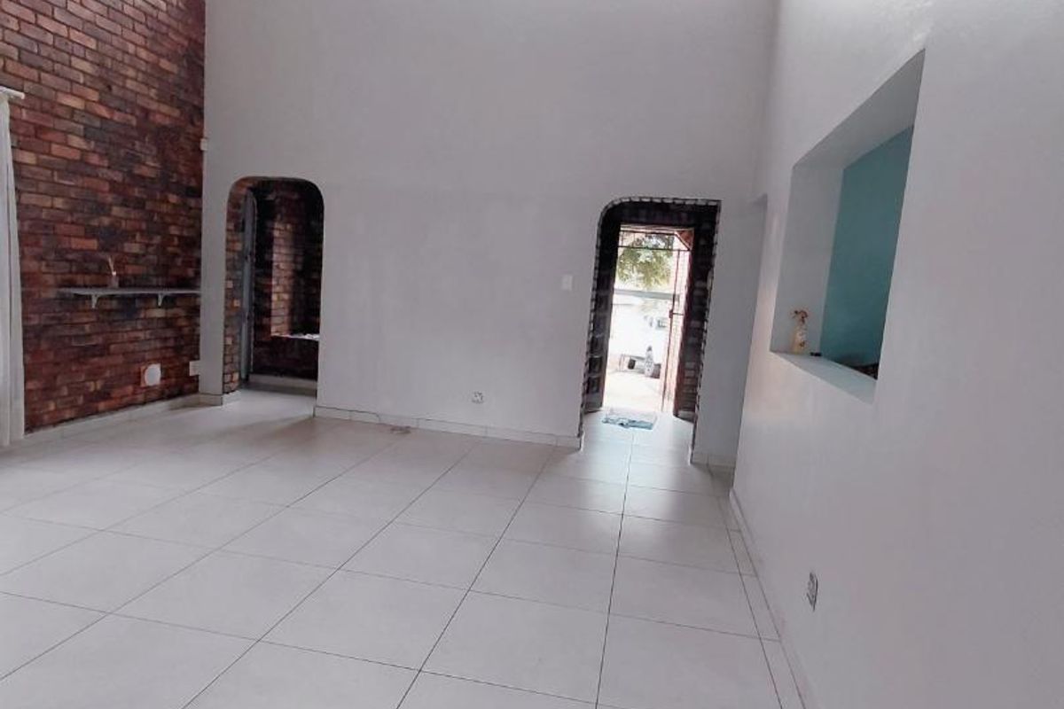 Spacious living area featuring high timber ceilings, exposed brick feature wall, clerestory windows for natural light, and neutral tiled flooring throughout.