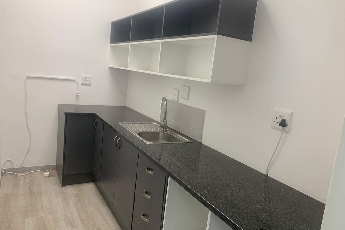 A modern office kitchenette featuring dark cabinetry, a stone-look countertop, stainless steel sink, and overhead storage shelving.