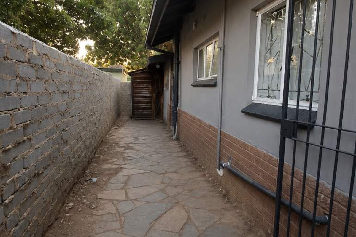A narrow side passage featuring stone paving, a brick boundary wall, and security gates attached to the exterior of the house.