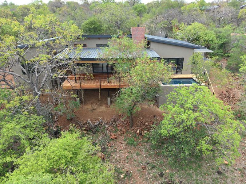 Aerial view of a modern bushveld home featuring a raised timber deck, integrated plunge pool, and a prominent brick chimney set in natural surroundings.