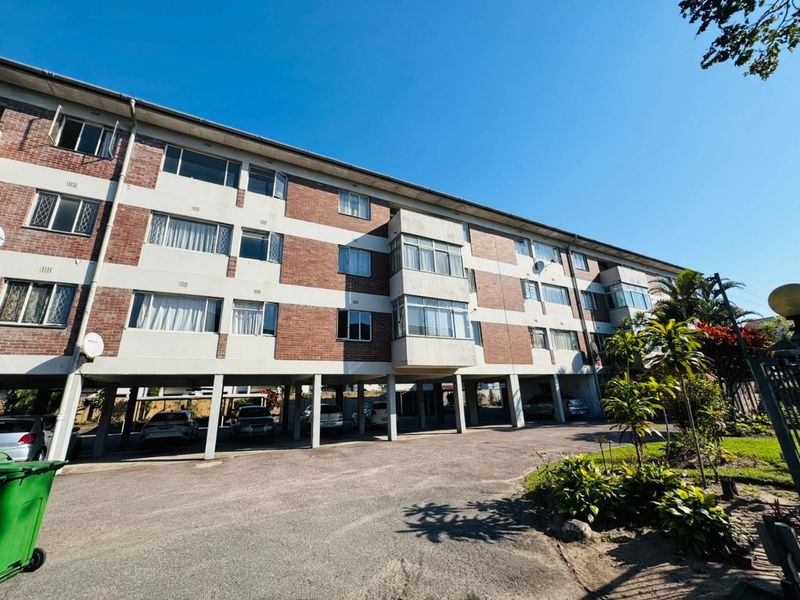 Exterior view of a multi-story apartment building featuring a brick facade, stilted ground-level parking area, and established surrounding landscaping.