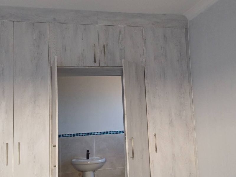 A bedroom featuring extensive built-in cupboards with a light wood-grain finish, providing direct access to an en-suite bathroom with a pedestal basin.