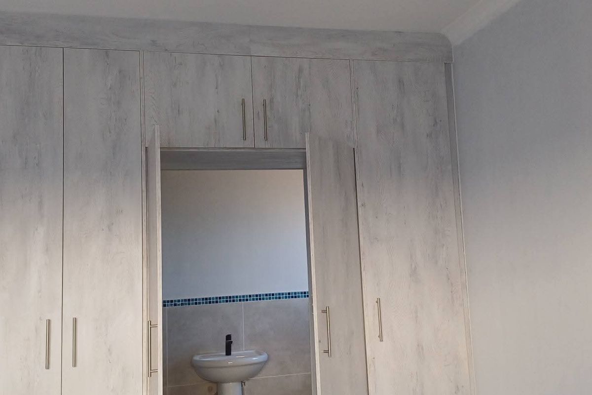 A bedroom featuring extensive built-in cupboards with a light wood-grain finish, providing direct access to an en-suite bathroom with a pedestal basin.
