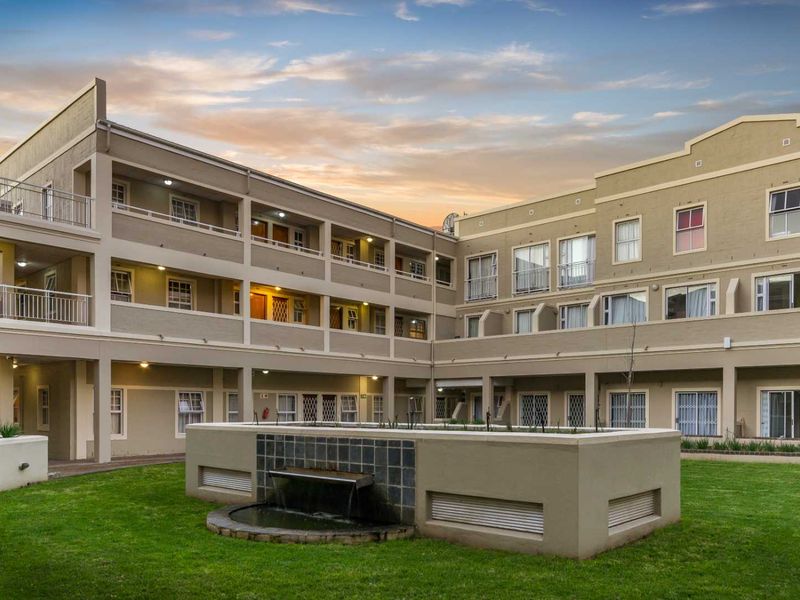 A well-maintained apartment complex exterior featuring a central courtyard with a water feature, landscaped lawn, and multi-level residential units with balconies.
