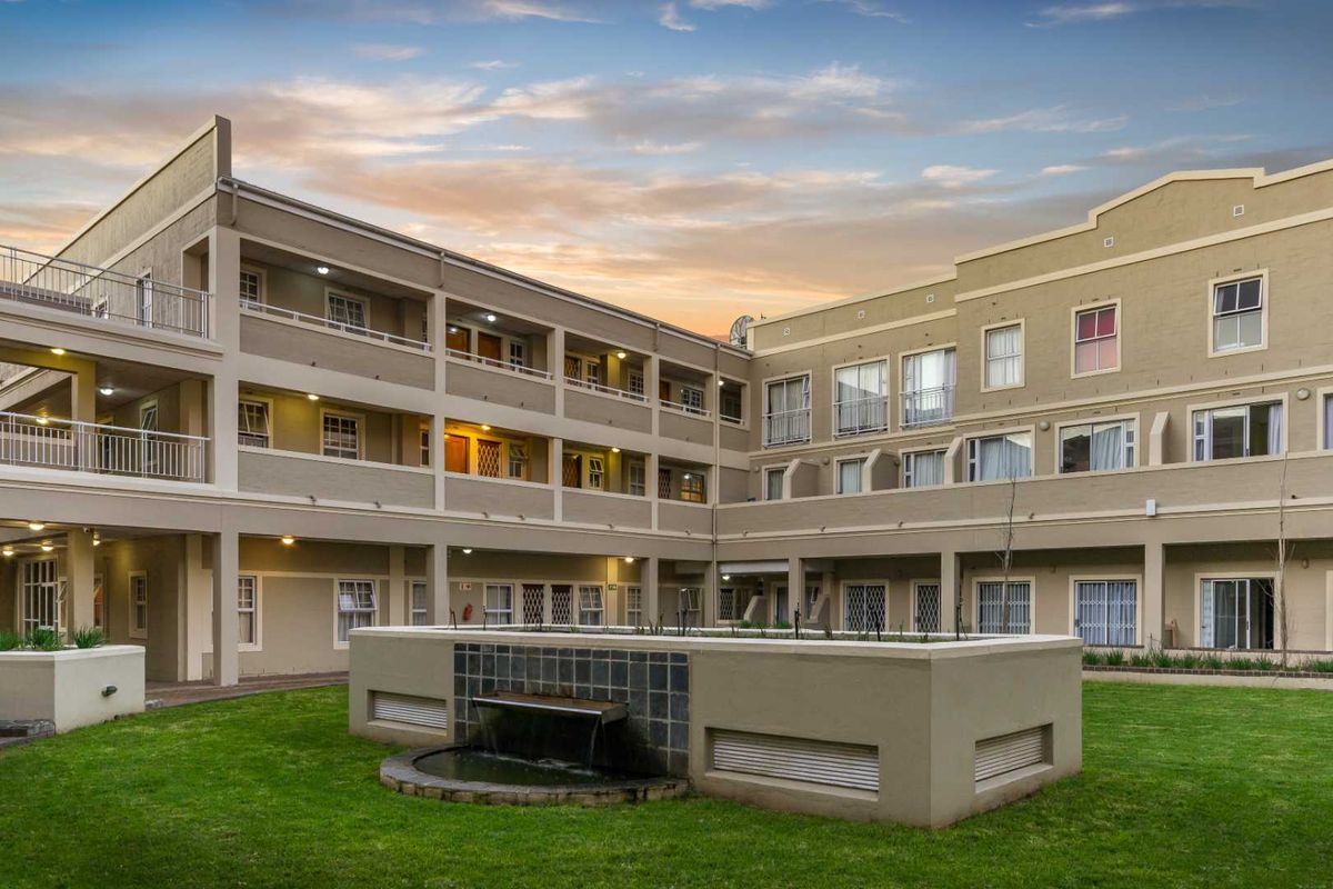 A well-maintained apartment complex exterior featuring a central courtyard with a water feature, landscaped lawn, and multi-level residential units with balconies.