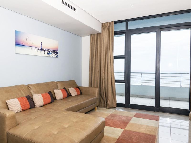 A bright living room featuring tiled floors, ducted air conditioning, and large sliding glass doors providing access to a balcony with ocean views.