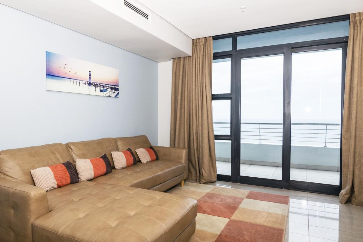 A bright living room featuring tiled floors, ducted air conditioning, and large sliding glass doors providing access to a balcony with ocean views.