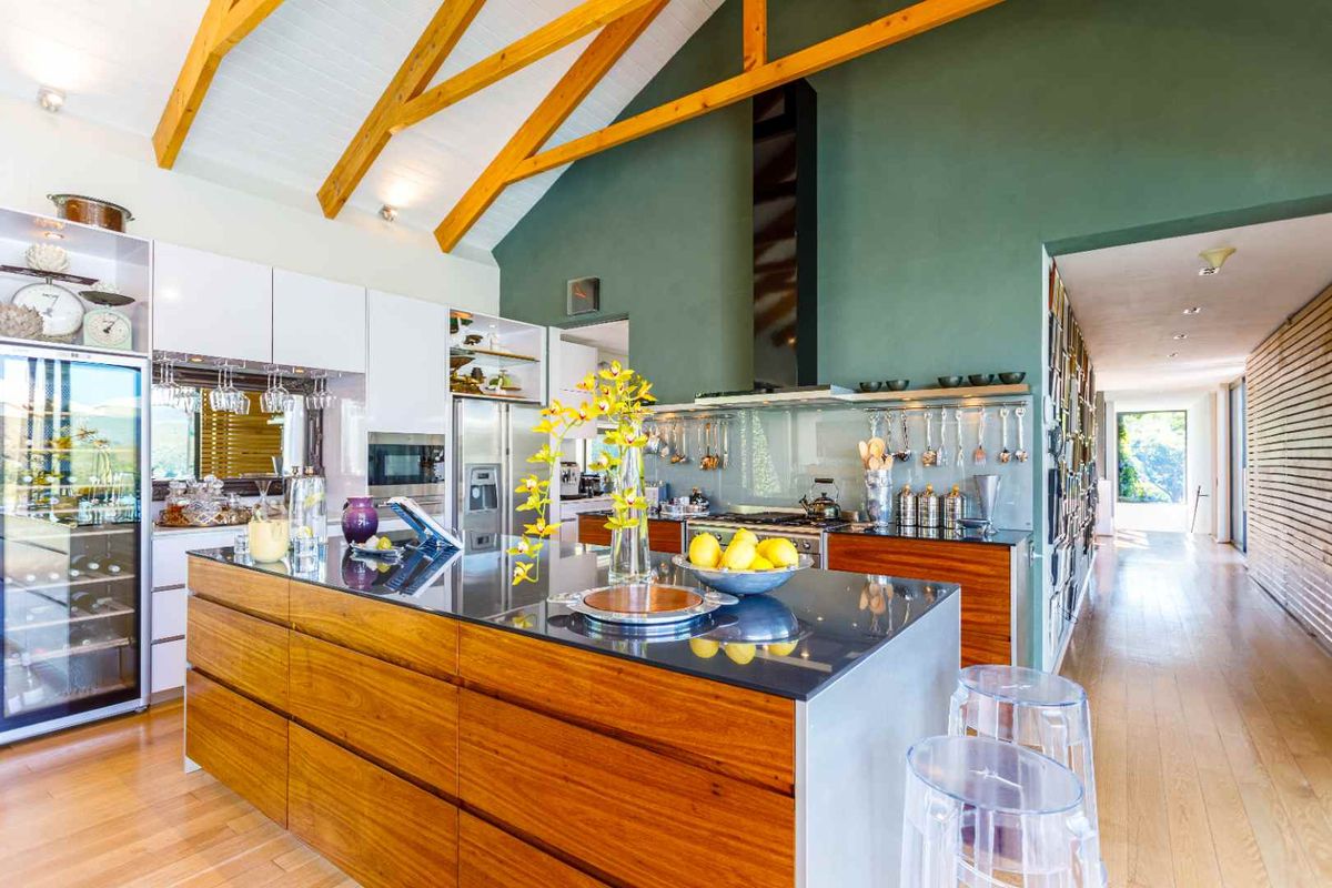 Modern kitchen featuring a large central island with stone countertop, timber cabinetry, high vaulted ceilings with exposed beams, and a glass splashback.