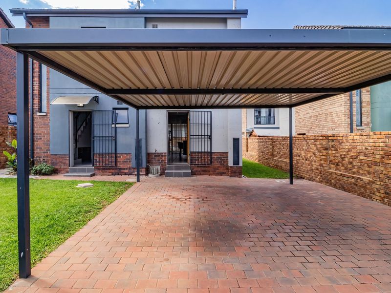 Rear exterior view featuring a paved driveway with a modern carport, brick boundary walls, and a well-maintained lawn.
