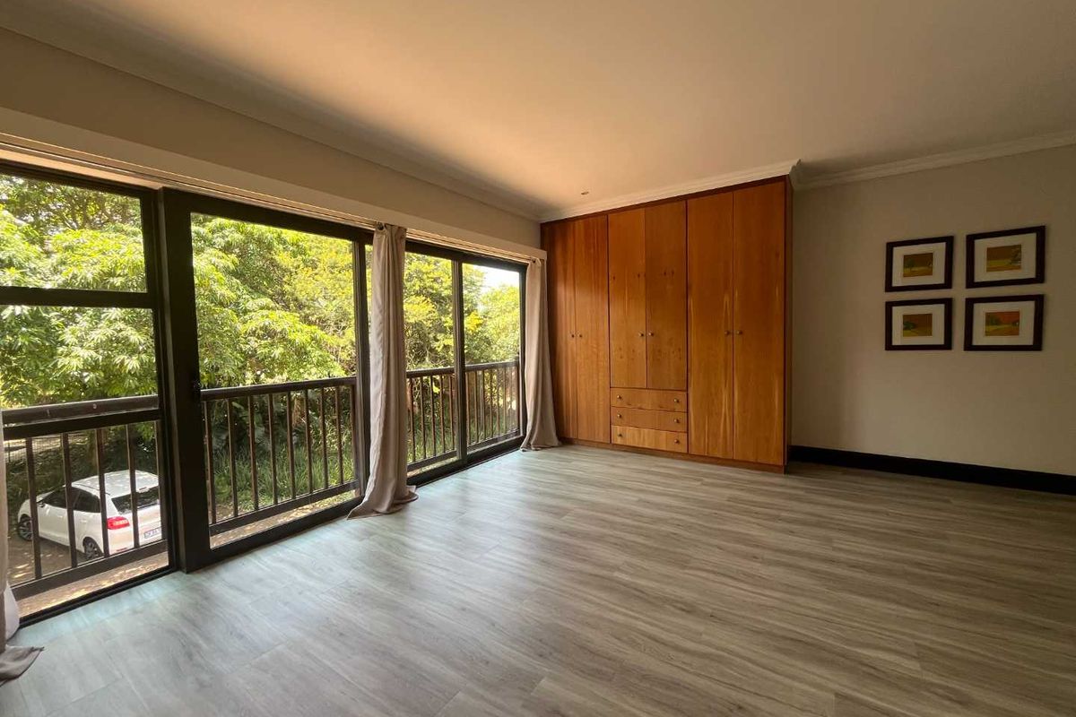 A bright bedroom featuring wood-look laminate flooring, built-in wooden cabinetry, and large sliding glass doors opening to a balcony with leafy views.
