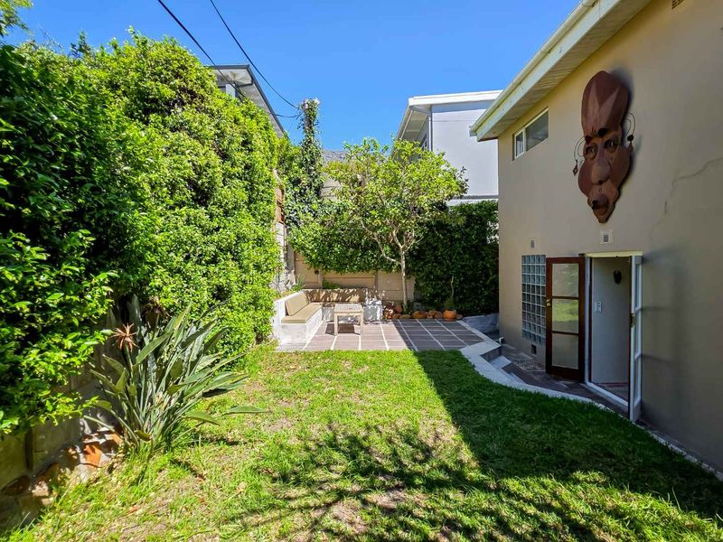 A private rear garden featuring a paved patio area with built-in bench seating, lush greenery, and direct access to the home via french doors.