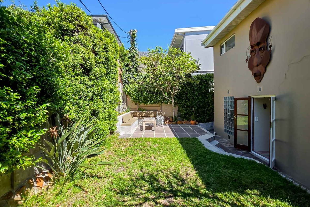 A private rear garden featuring a paved patio area with built-in bench seating, lush greenery, and direct access to the home via french doors.