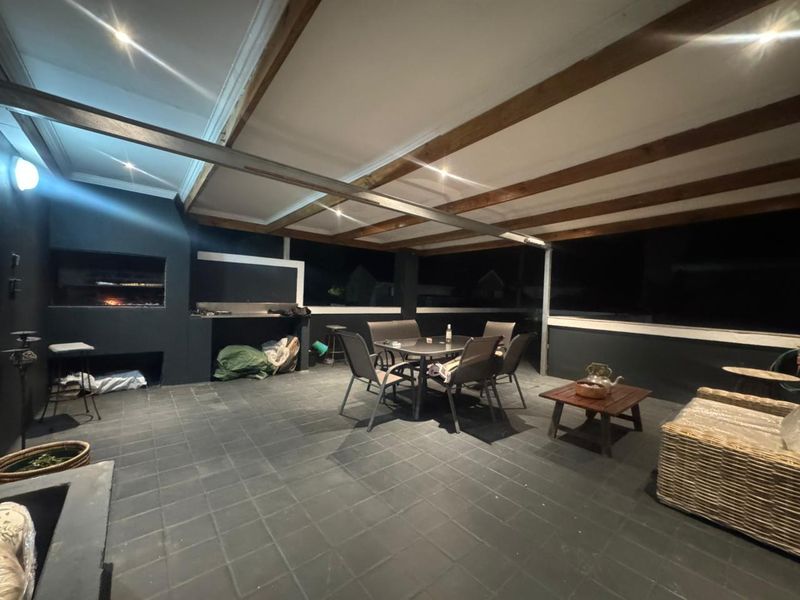 A spacious covered patio featuring a built-in braai, exposed timber ceiling beams, and dark tiled flooring, ideal for outdoor entertaining.