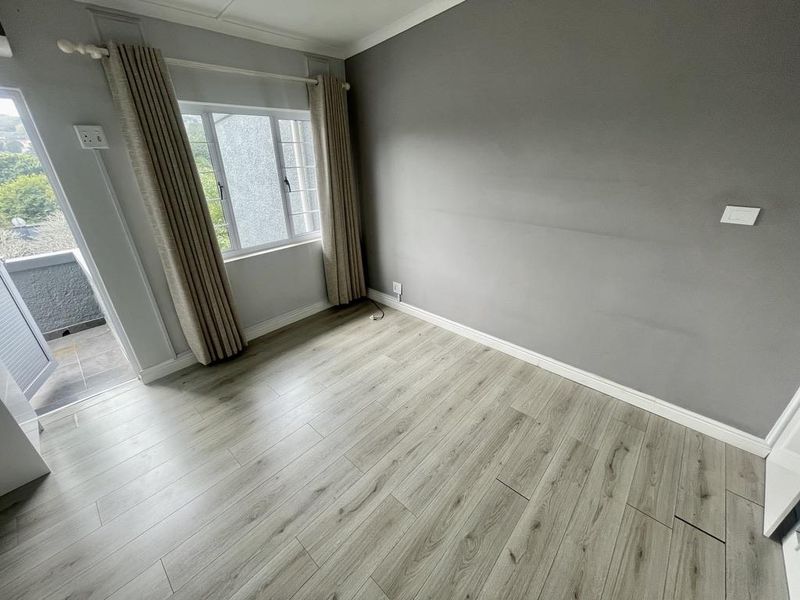 A bright, empty bedroom featuring modern laminate flooring, neutral grey walls, and direct access to a private balcony through a glass door.