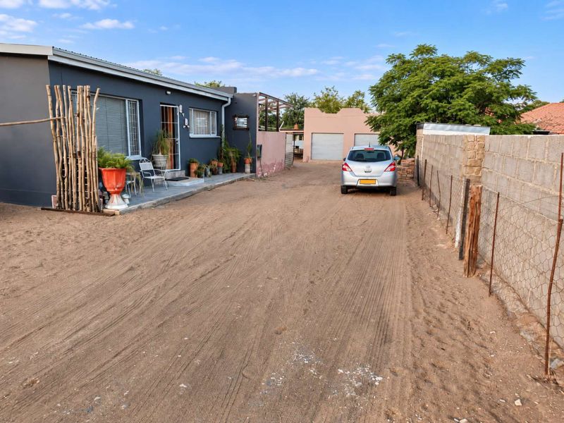 Front exterior view showing a house with a patio, a double garage, and a large unpaved driveway area enclosed by a boundary wall.