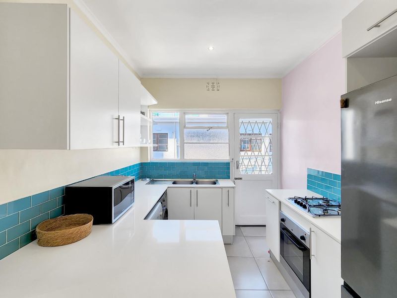 A bright, functional kitchen featuring white cabinetry, quartz countertops, a gas hob, built-in oven, and vibrant blue tiled backsplash with direct exterior access.