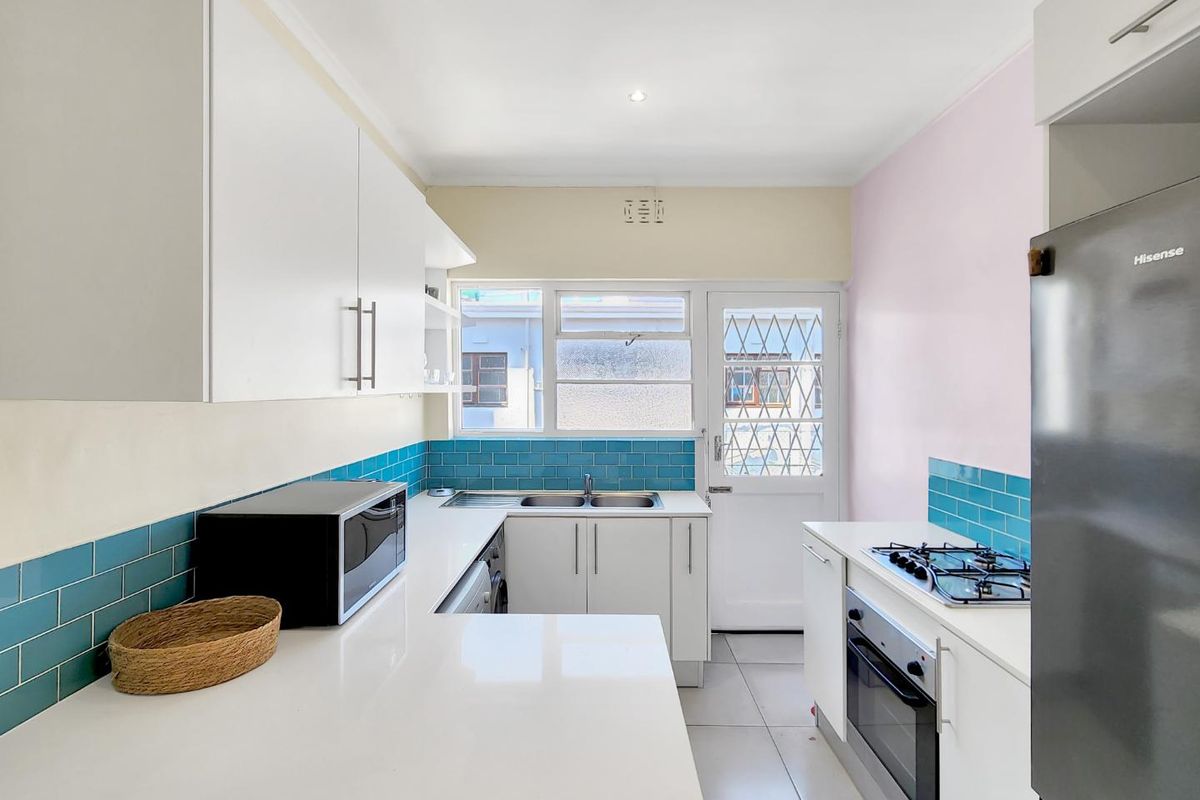 A bright, functional kitchen featuring white cabinetry, quartz countertops, a gas hob, built-in oven, and vibrant blue tiled backsplash with direct exterior access.