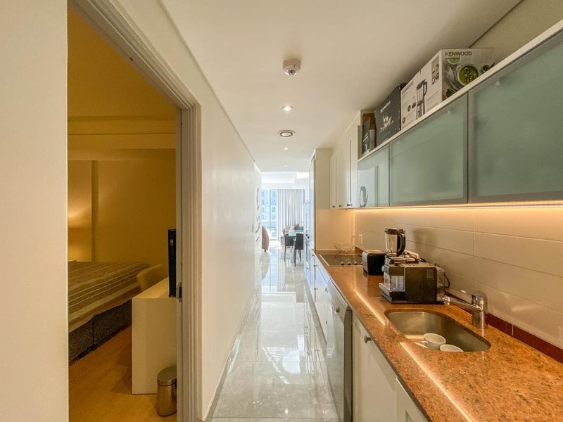 A compact, modern galley-style kitchen featuring granite countertops, integrated cabinetry, and under-cabinet lighting, leading into an open-plan living area.