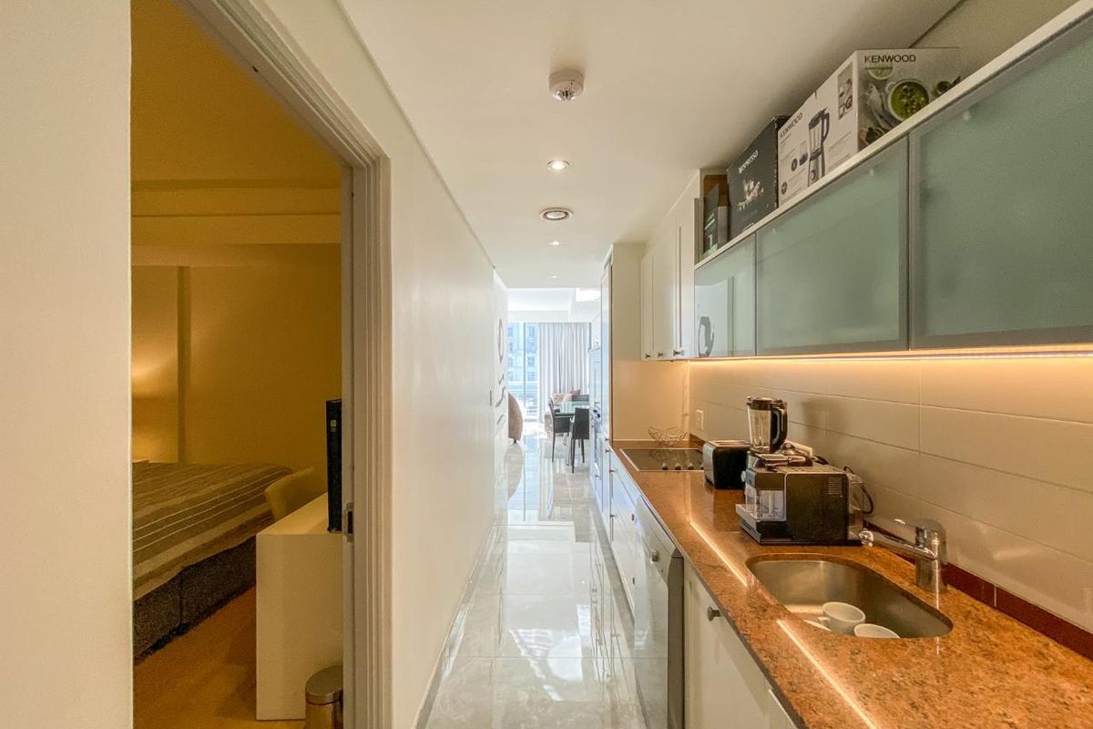 A compact, modern galley-style kitchen featuring granite countertops, integrated cabinetry, and under-cabinet lighting, leading into an open-plan living area.