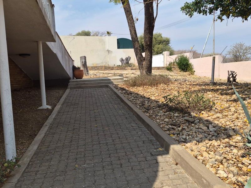 A paved walkway leads through the rear garden area, featuring a brick retaining wall and concrete support columns under an elevated section of the house.