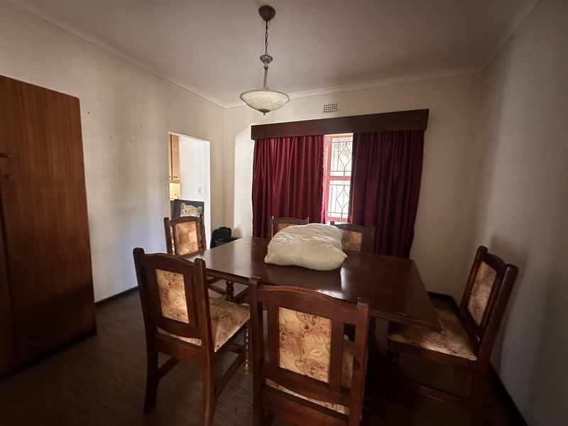 A dining room featuring laminate flooring, a central light fixture, and a window with security bars, providing access to an adjacent kitchen area.
