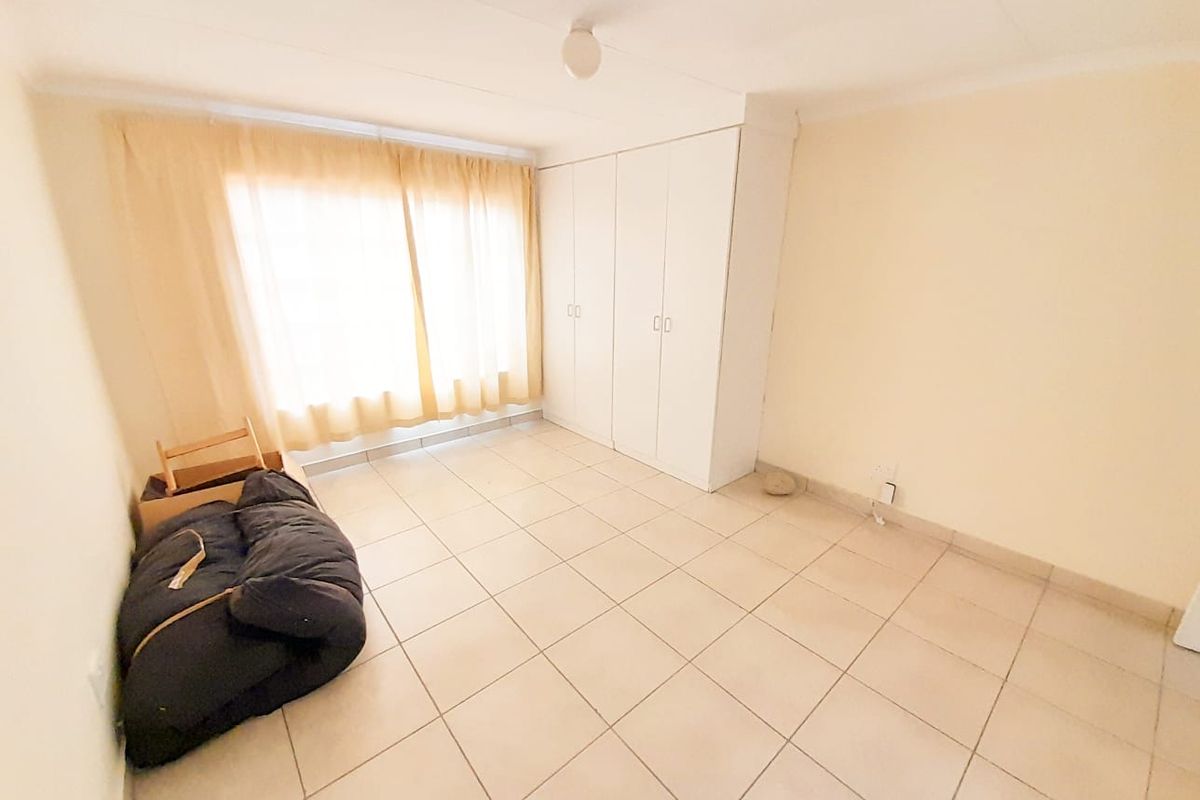A bright bedroom featuring neutral tiled flooring, built-in cupboards, and large windows with curtains, offering a clean and functional living space.