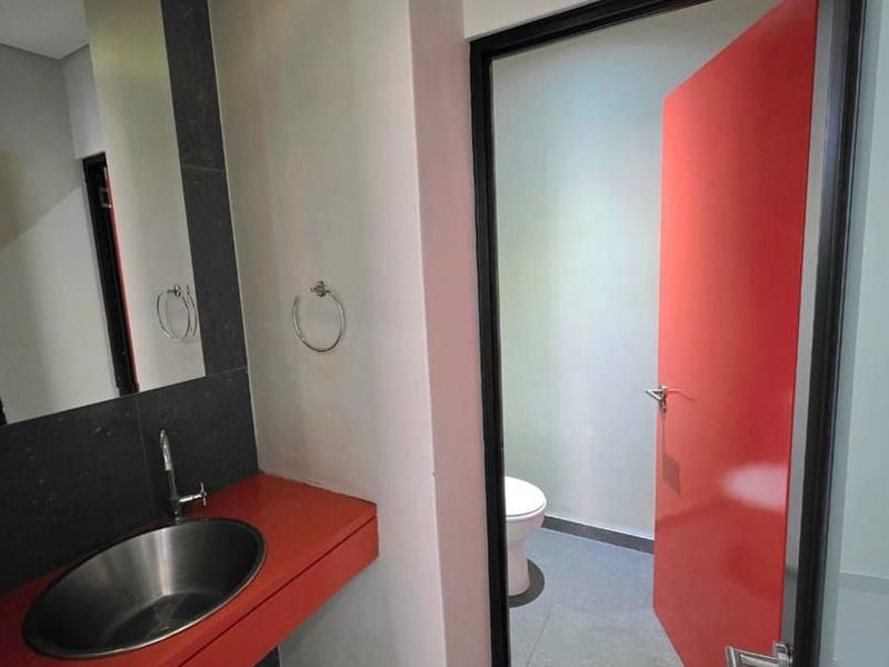 Modern office bathroom featuring a sleek vessel sink, tiled vanity, and a separate toilet cubicle with a bold red door.