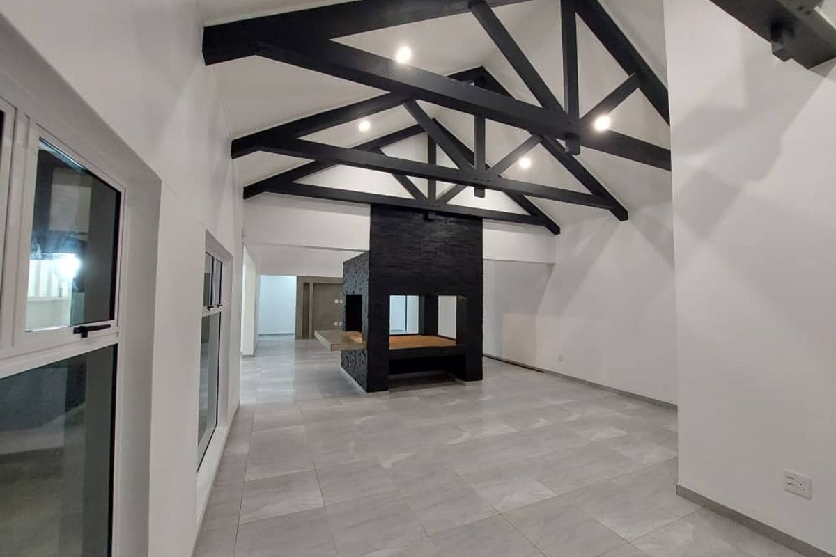 Spacious living area featuring high vaulted ceilings with exposed black timber trusses, modern tiled flooring, and a striking central built-in fireplace.