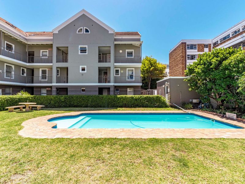 Communal swimming pool area featuring a paved surround, well-maintained lawn, and established garden hedging, set against the apartment building facade.