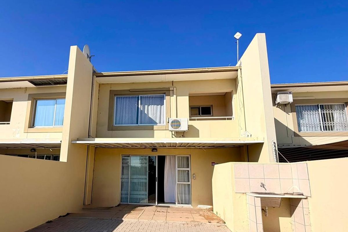 Rear exterior view of the townhouse featuring a paved patio area, built-in braai, sliding door access, and an upper-level balcony.