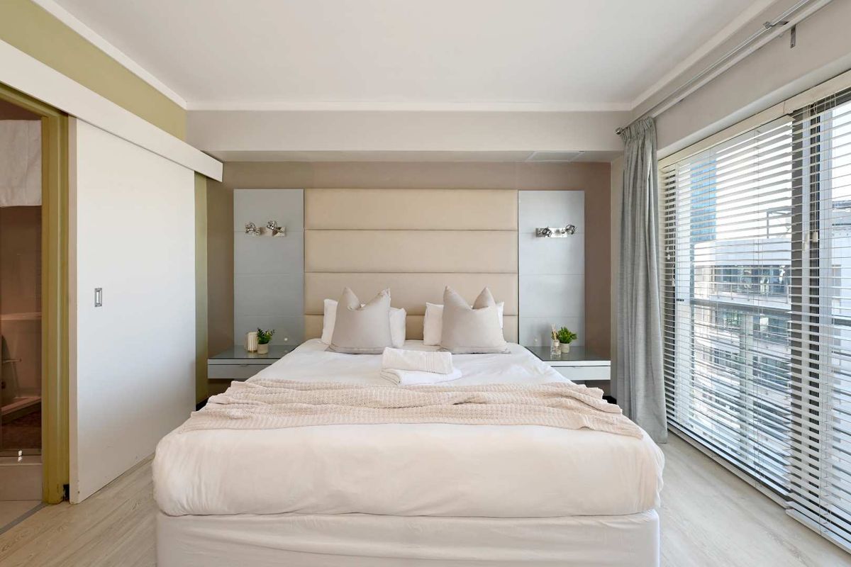 A well-maintained bedroom featuring a custom upholstered headboard, integrated bedside tables, wall-mounted lighting, and large windows with venetian blinds.