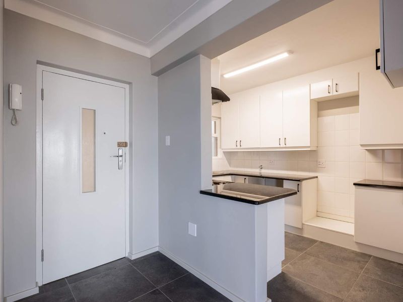 A clean, modern kitchen featuring white cabinetry, dark countertops, tiled backsplash, and grey floor tiles, with an adjacent entrance door and intercom system.
